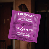 A large purple packet of LifeStyles "Snugger Fit" Condoms partially obscures a naked person with long hair standing in a dimly-lit room with reddish walls and a door in the background. The skin-tight fit packet, prominently centered in the image, adds an intriguing focus to the scene.